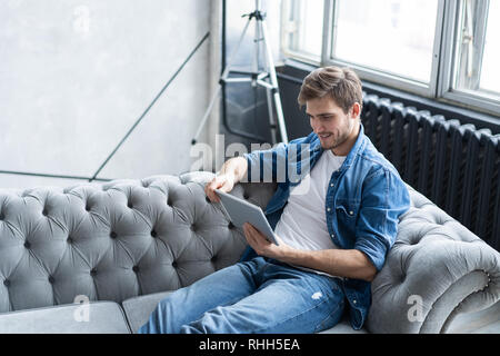 Schöner Mann ist mit einem digitalen Tablet und lächelnd, während auf der Couch zu Hause ausruhen. Stockfoto