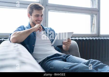 Schöner Mann ist mit einem digitalen Tablet und lächelnd, während auf der Couch zu Hause ausruhen. Stockfoto