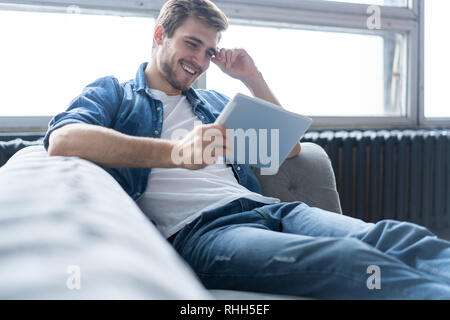 Schöner Mann ist mit einem digitalen Tablet und lächelnd, während auf der Couch zu Hause ausruhen. Stockfoto