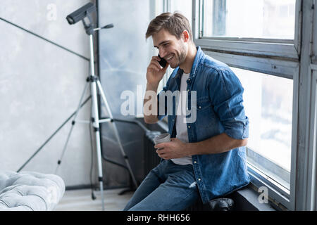 Lächelnden jungen Mann Gespräch am Handy, am Fenster zu Hause suchen. Stockfoto