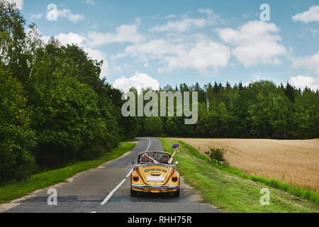 Golden retro Cabriolet mit ein paar frisch verheiratet, auf der leeren Straße zwischen Feld und Wald Stockfoto