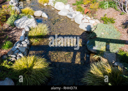 Schatten von einer Person, die auf einer Brücke über einen kleinen Bach von Wasser Stockfoto