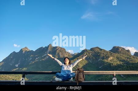 Asiatische Frau Reisende sitzen und die Arme in die Luft an View Point Terrasse an Landschaft, Blick auf die Berge mit Cloud und blauer Himmel im sonnigen Tag an Fores Stockfoto