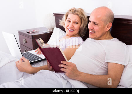 Freudige reifer Mann und Frau im Bett mit Laptop und Buch. Fokus auf den Menschen Stockfoto