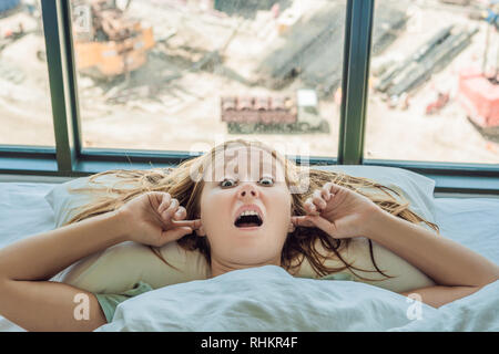 Junge Frau liegen auf einem Bett bedeckte ihre Ohren, weil der Lärm. Im Fenster nach dem Bett können Sie den Bau eines neuen Hauses Stockfoto