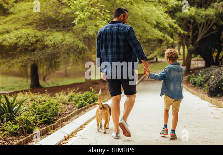 Hintere Ansicht von Vater und Sohn mit Hund wandern in einem Park. Mann hält eine Hand auf seinen kleinen Jungen gehen und sprechen, draußen im Park. Stockfoto