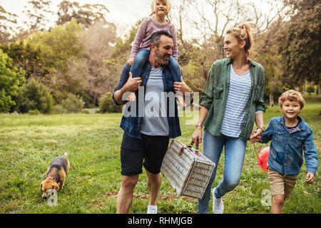 Glückliche Familie mit Hund auf dem Weg zur Laube im Garten. Mann, der seine Tochter auf der Schulter mit Frau mit Sohn und mit einer Hand Stockfoto