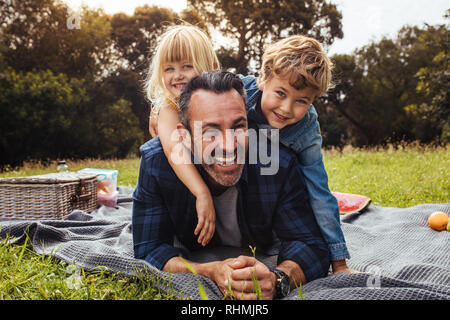 Glückliche Menschen mit Kindern bei einem Picknick im Park neben einem Picknickkorb. Kinder liegen auf dem Rücken ihres Vaters. Stockfoto