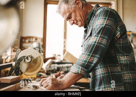 Senior Zimmermann Notizen in seinem Buch in der Werkstatt. Reife männliche Tischler seine Arbeit Planung Zimmerei. Stockfoto