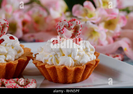 Selbstgemachte Valentine süße kleine Kuchen mit Sahne Zucker Cap mit roten Herzen und rosa Blumen Hintergrund Stockfoto