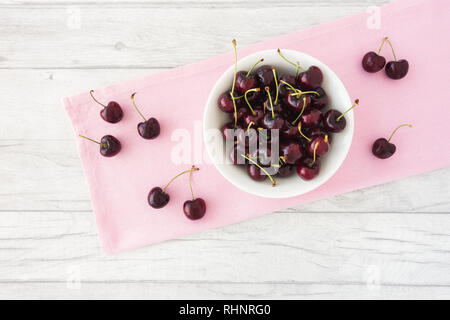 Kirschen in ein weißes Porzellan Schüssel und auf einem rosa Stoff auf einem weißen Laufbelag Hintergrund verstreut. Ansicht von oben. Stockfoto