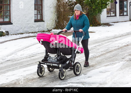 Junge Frau drückt double Buggy entlang der schneebedeckten Straße an einem kalten Wintertag in Glencoe Dorf, Highlands, Schottland im Winter Stockfoto