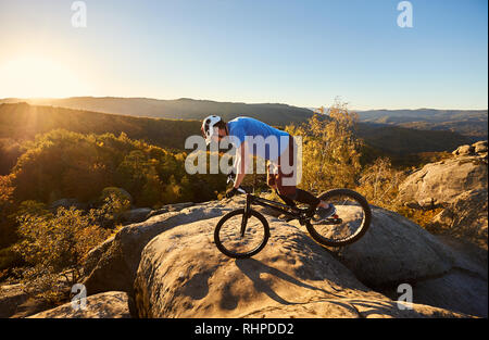 Mutige Radfahrer stehen auf Vorderrad auf Trial Fahrrad. Professionelle Sportler biker Balancing am Rande des großen Felsen auf dem Gipfel des Berges bei Sonnenuntergang. Konzept der Extreme Sport Stockfoto