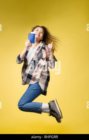 Mädchenhafte, funky, Glück, Traum, Spaß, Freude, Sommer Konzept. Sehr aufgeregt, fröhlich cute Asian teen springt mit Reisepass gelben Hintergrund. Stockfoto