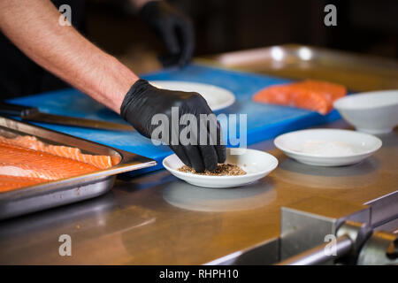 Thema Kochen ist ein Beruf des Kochens. Nahaufnahme der Hand ein kaukasischer Mann in einem Restaurant Küche vorbereiten rot Fischfilets Lachs Fleisch in schwarz l Stockfoto