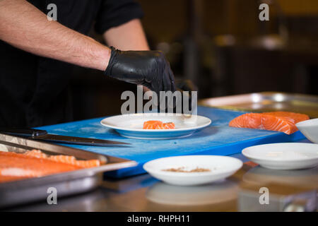 Thema Kochen ist ein Beruf des Kochens. Nahaufnahme der Hand ein kaukasischer Mann in einem Restaurant Küche vorbereiten rot Fischfilets Lachs Fleisch in schwarz l Stockfoto