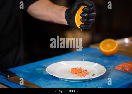 Thema Kochen ist ein Beruf des Kochens. Nahaufnahme der Hand ein kaukasischer Mann in einem Restaurant Küche vorbereiten rot Fischfilets Lachs Fleisch in schwarz l Stockfoto