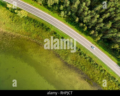 Luftbild von Oben nach Unten Blick auf zweispurigen Straße mit Pinienwäldern auf der einen Seite und Tiefe grüne See auf der anderen. Schönen Sommer Landschaft in Litauen. Stockfoto