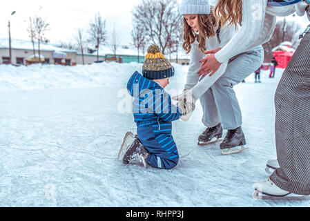 Eine junge Mutter mit Freundin hilft stand von Hand kleine Junge 2-4 Jahre altes Kind. Im Winter, Eisbahn in der Stadt. Rest frische Luft. Die ersten Schritte auf den Skates Stockfoto