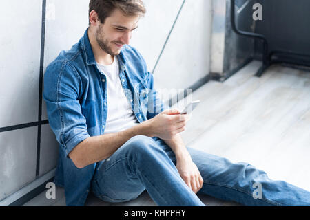 Junge lächelnde Mann sitzt auf dem Boden, während Sie ihr Smartphone gegen graue Wand. Stockfoto