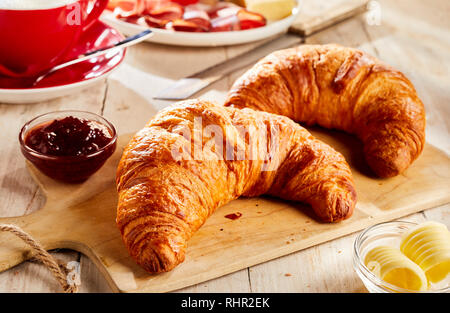 Zwei frisch gebackene Croissants auf Holz Schneidebrett, serviert mit Marmelade und Butter in kleinen Glasschalen Stockfoto