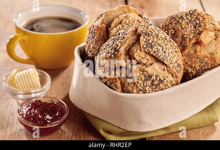 Brötchen frisch gebackenen knusprigen Schwarzbrot serviert in Stofftasche, mit Butter, Schale, Marmelade und Tasse Kaffee auf Holztisch, in enger gesehen - bis Stockfoto