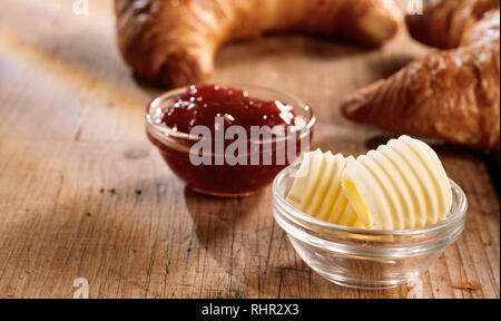 Frische Locken von Butter und Marmelade im Glas Schalen zum Frühstück mit Croissants, auf zerkratzte Holz Tisch serviert und gesehen in Nahaufnahme Stockfoto