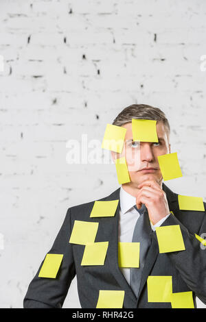 Nachdenklich reife Geschäftsmann mit leeren gelben Klebenotizen auf Gesicht und Klage gegen Wand im Büro Stockfoto