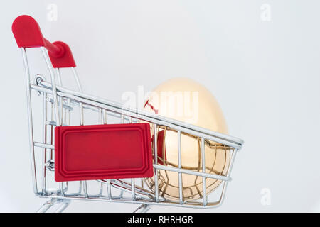 Ein goldenes Ei aus Metall in einem Supermarkt Lebensmittel Warenkorb auf einen hellgrauen Hintergrund. Das Konzept einer einzigartigen Angebot für den Kunden. Kopieren Sie Platz. Stockfoto