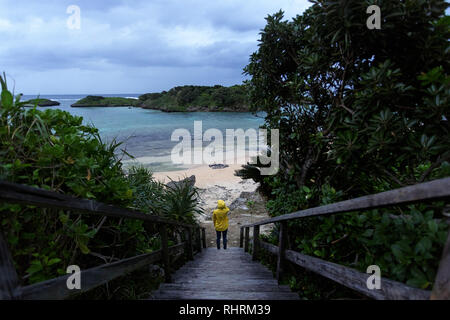 Frau in gelb Regenjacke stehen auf hölzernen Promenade zum schönen Sandstrand von tropischen Strand bei stürmischem Wetter, Japan Stockfoto