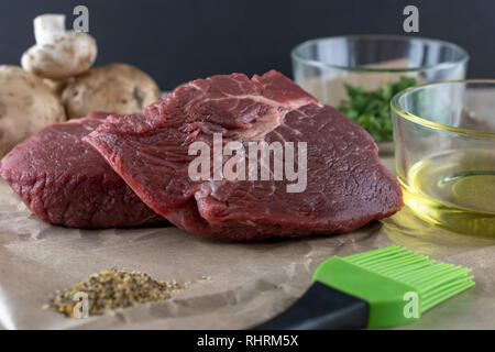 Rohe Steaks prepped für das Kochen. Gewürze im Vordergrund, Pilze im Hintergrund. Schwarzen Hintergrund. 2 Glasschüsseln mit Olivenöl und Petersilie Stockfoto