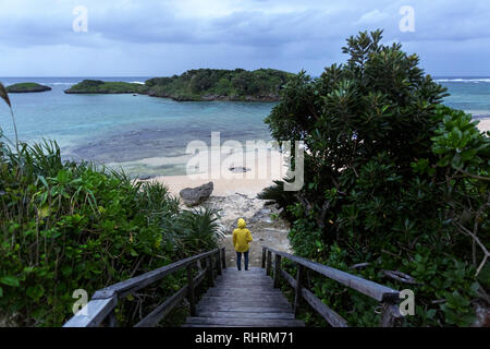 Frau in gelb Regenjacke stehen auf hölzernen Promenade zum schönen Sandstrand von tropischen Strand bei stürmischem Wetter, Japan Stockfoto