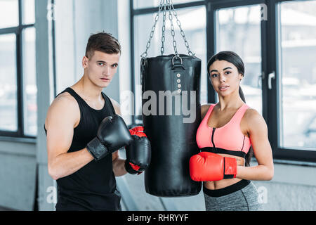 Männliche und weibliche Boxer in Boxhandschuhe in der Nähe von Boxsack und Kamera in der Turnhalle stehen Stockfoto