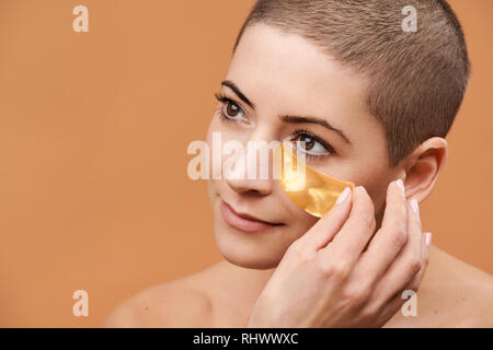 Lächelnd Mitte 30 Frau Anwendung gold Collagen Augenmaske. Foto von attraktiven kaukasische Frau mit Kopf rasiert und gesunde Haut isoliert auf beige backgro Stockfoto