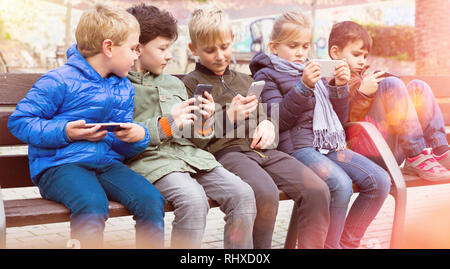 Gruppe von jugendlichen Kindern sitzt auf der Bank im Freien, in ihre Telefone süchtig Stockfoto