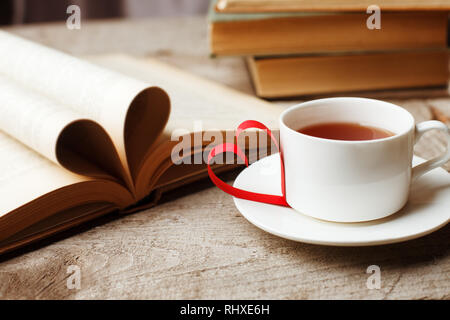 Liebe für Bücher, Lesen. Stapel Bücher auf dem Holztisch. Buch mit gewellten Blätter in der Form eines Herzens öffnen, Tasse Tee. Bibliothek, Bildung, Leis Stockfoto