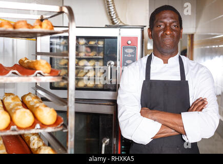Zuversichtlich Baker in schwarze Schürze mit Waffen in der Nähe von professionellen Brotbackofen in kleine Bäckerei gekreuzt Stockfoto