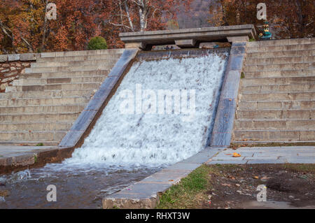 Ein künstlicher Wasserfall in einem öffentlichen Park. Bäume mit gelben Blätter im Herbst. Stockfoto