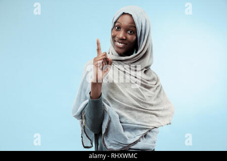 Die schöne junge schwarze afrikanische muslimische Mädchen mit grauen Hijab im Blue Studio. Sie stehen mit einem glücklichen Lächeln auf ihrem Gesicht. Die menschlichen Gefühle, Mimik Konzept. Trendige Farben Stockfoto