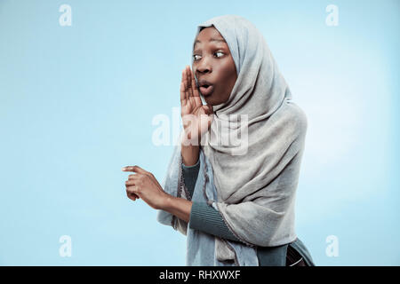 Geheimnis, Klatsch-Konzept. Junge afrikanische Frau flüstert ein Geheimnis hinter ihrer Hand. Die Frau isoliert am trendigen Blau studio Hintergrund. Junge emotionale Frau. Menschliche Gefühle, Mimik Konzept. Stockfoto