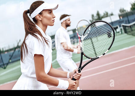 Junge Frau und Mann in Weiß Sportswear stehen auf dem Hof mit Schläger in der Hand voll und ganz auf das Spiel konzentriert Stockfoto