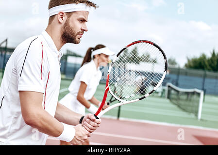 Junge Frau und Mann in Weiß Sportswear stehen auf dem Hof mit Schläger in der Hand voll und ganz auf das Spiel konzentriert Stockfoto