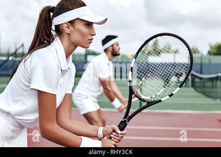 Junge Frau und Mann in Weiß Sportswear stehen auf dem Hof mit Schläger in der Hand voll und ganz auf das Spiel konzentriert Stockfoto