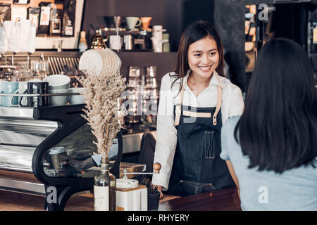 Asiatische Frau barista Schürze tragen Jean holding Tasse Kaffee serviert, um Kunden an Theke mit Smile Emotion, Cafe Restaurant Service Konzept, Inhaber smal Stockfoto