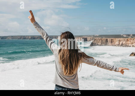 Rückansicht eines Mädchen bewundern die schöne Aussicht auf den Atlantischen Ozean und die Landschaft und ihre Hände zeigen, wie froh sie ist. Stockfoto