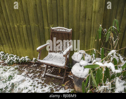 Holz garten Stuhl mit Schnee neben einem grünen Zaun und Behälter mit Schnee auf ihre Deckel abgedeckt. Schnee auf die Büsche und Masse. Stockfoto