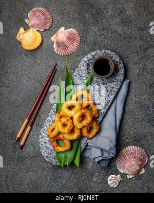 Frittierte Tintenfischringe auf der Steinplatte mit tropischen Blättern verziert. Asiatische Lebensmittel Konzept, Sojasauce und Essstäbchen Stockfoto