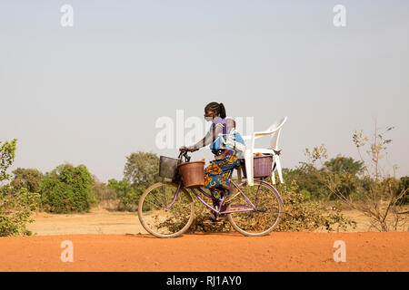 Samba Dorf, yako Provinz, Burkina Faso: Eine junge Frau trägt Ihr Baby auf den Rücken und Eimer mit einem Stuhl auf ihr Fahrrad. Stockfoto