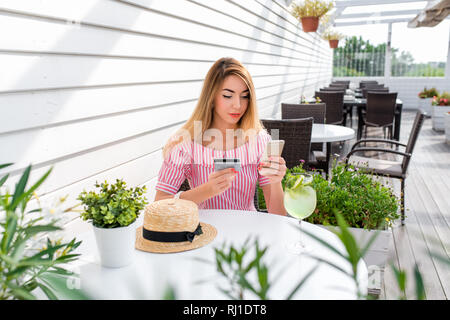 Schöne Mädchen in einem Café im Sommer in einem Kleid mit langen Haaren. In seiner Hand, eine Plastikkarte und Telefon. Eine Frau zahlt das Mittag- oder Abendessen mit einer Kreditkarte Stockfoto