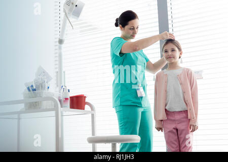 Krankenschwester Messhöhe von Mädchen gegen Fenster im Untersuchungsraum Stockfoto